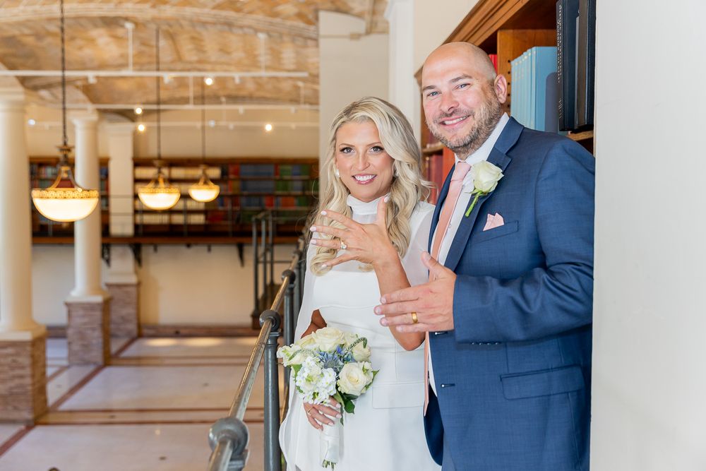 Smiling bride and groom pose together indoors, bride holding white bouquet, groom in blue suit with boutonniere.