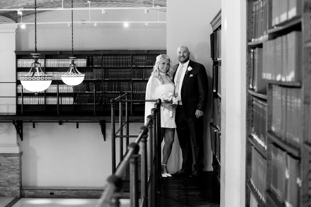 Black and white photo of a bride and groom standing on a balcony inside an elegant library venue.