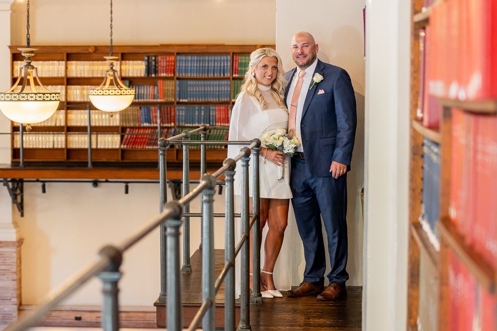 Bride and groom pose elegantly on a staircase landing in a library with bookshelves in the background.
