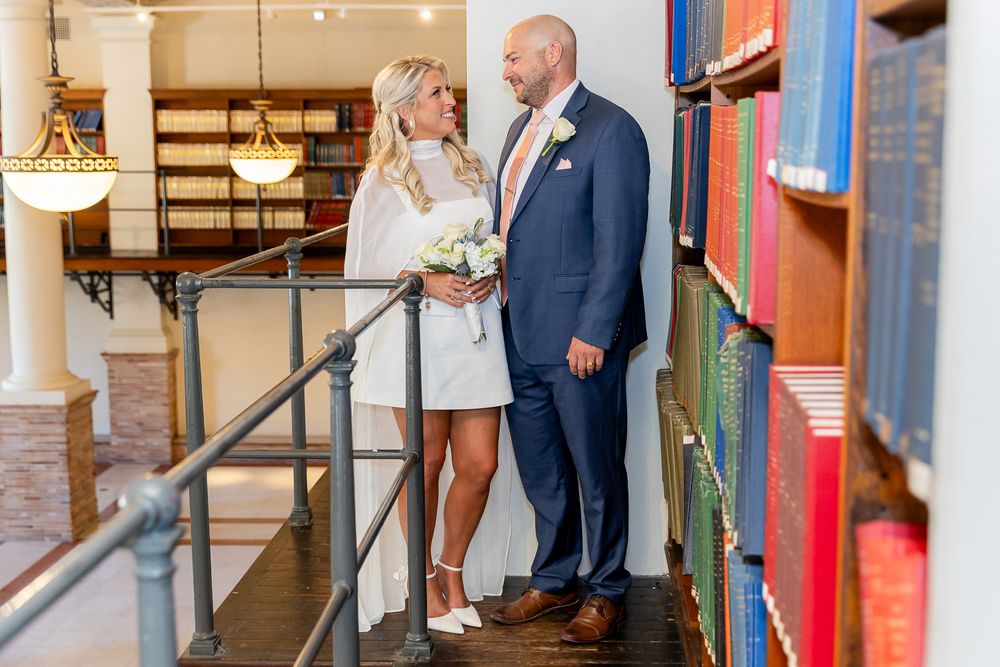 Bride and groom share a romantic moment on library stairs surrounded by colorful books on their wedding day.