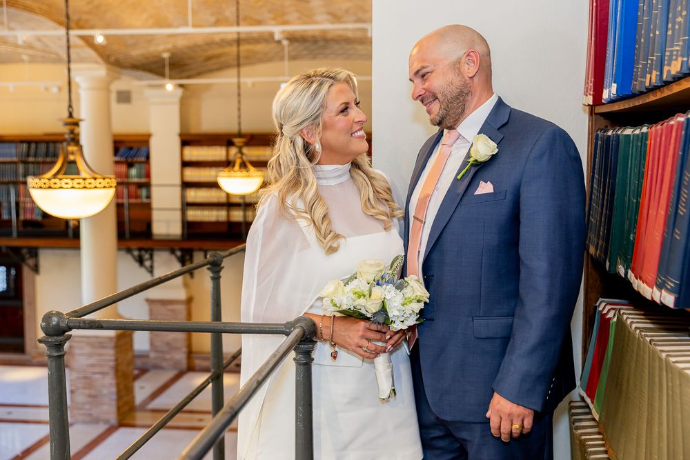 Bride and groom smiling at each other in a library setting, bride holding white bouquet, groom in navy suit with peach tie.