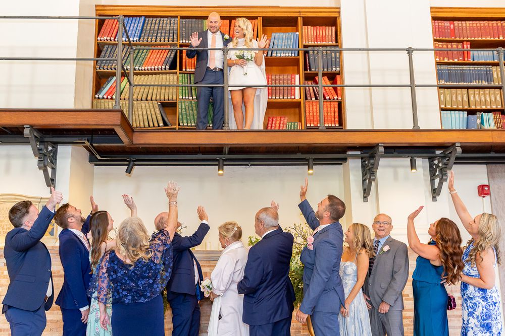 Bride and groom celebrate on a balcony surrounded by bookshelves as wedding guests cheer below.