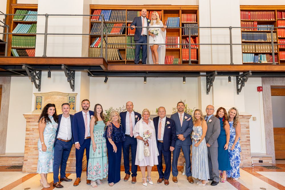 Wedding group photo with family posing in elegant library venue, bride and groom visible on balcony above.