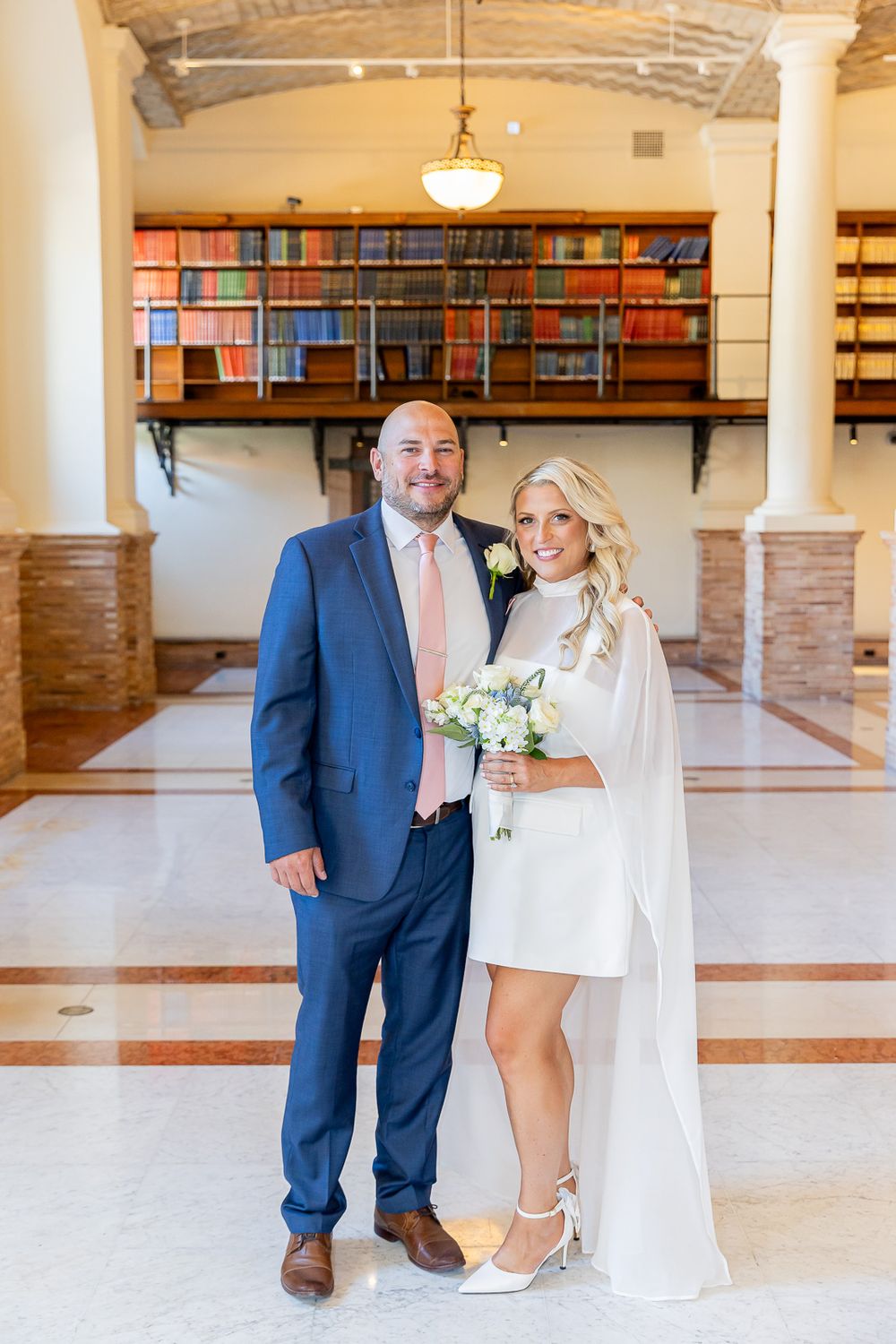 Couple posing in elegant library venue, groom in blue suit, bride in white mini dress with cape holding bouquet.