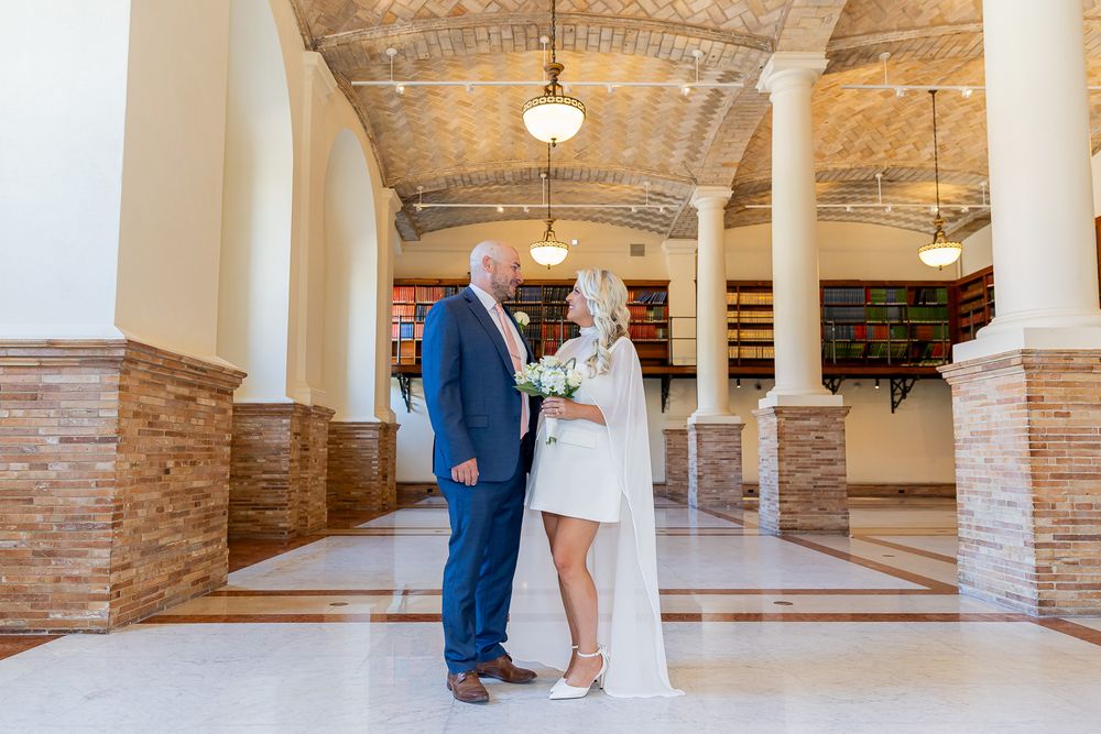 Couple in wedding attire standing in elegant hall with arched ceilings, bride holding bouquet, groom in blue suit.