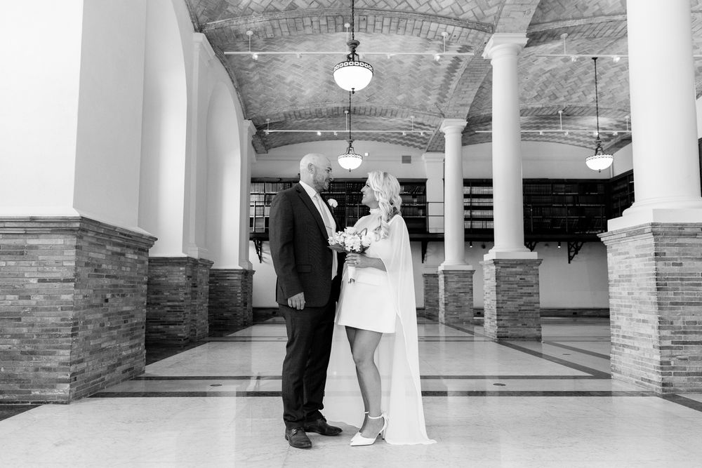 Black and white wedding photo of bride and groom standing in elegant columned hall with arched ceiling.