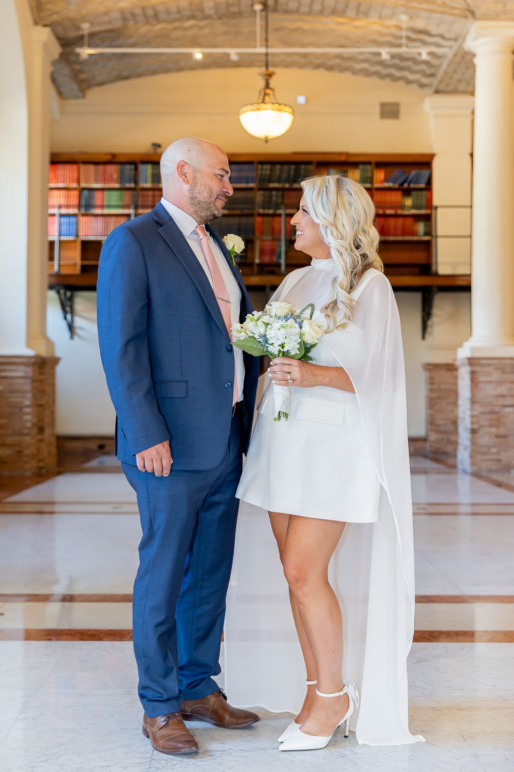Couple in wedding attire stand together in elegant hall, groom in blue suit, bride in white mini dress holding bouquet.