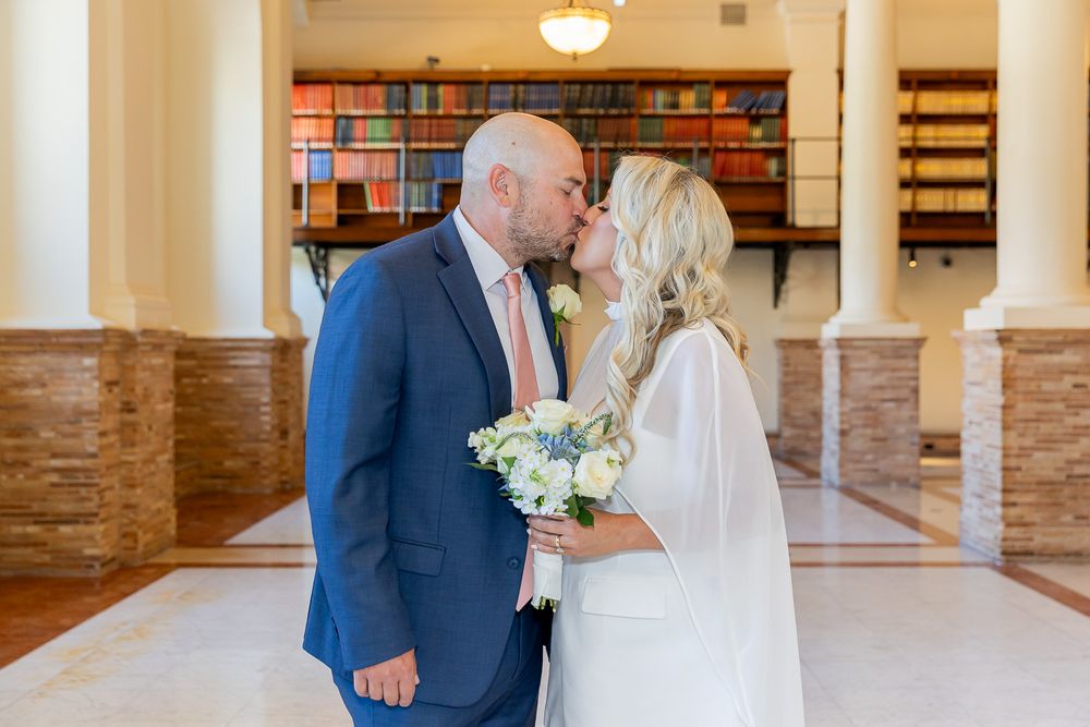 Bride and groom share a kiss in an elegant library venue, bride holding white floral bouquet in wedding dress.
