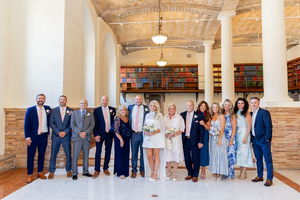 Wedding party group photo in a grand library hall with arched ceilings and bookshelves in background.