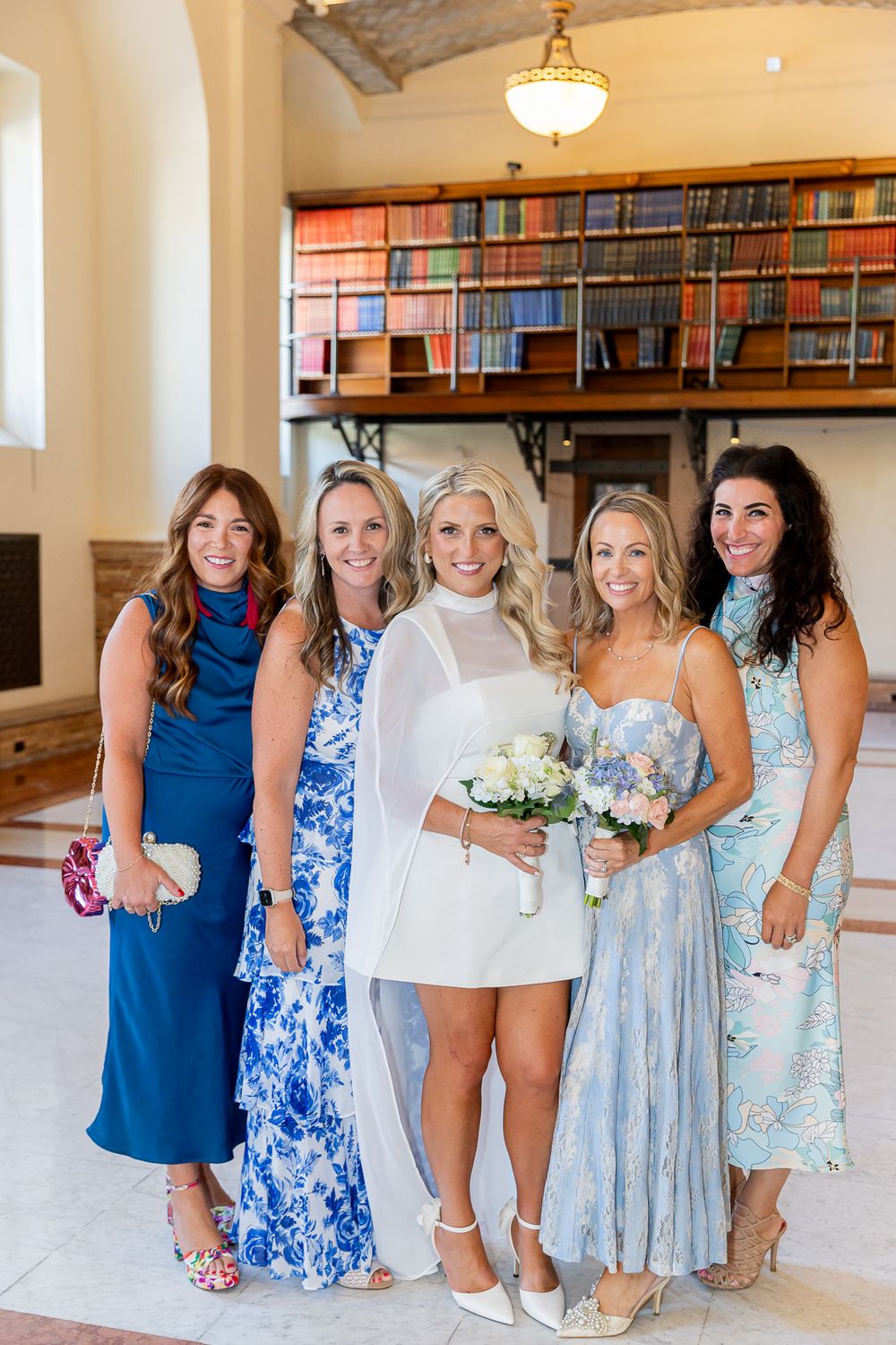 Bride in white mini dress holds bouquet, posing with four women in blue floral dresses inside elegant library venue.