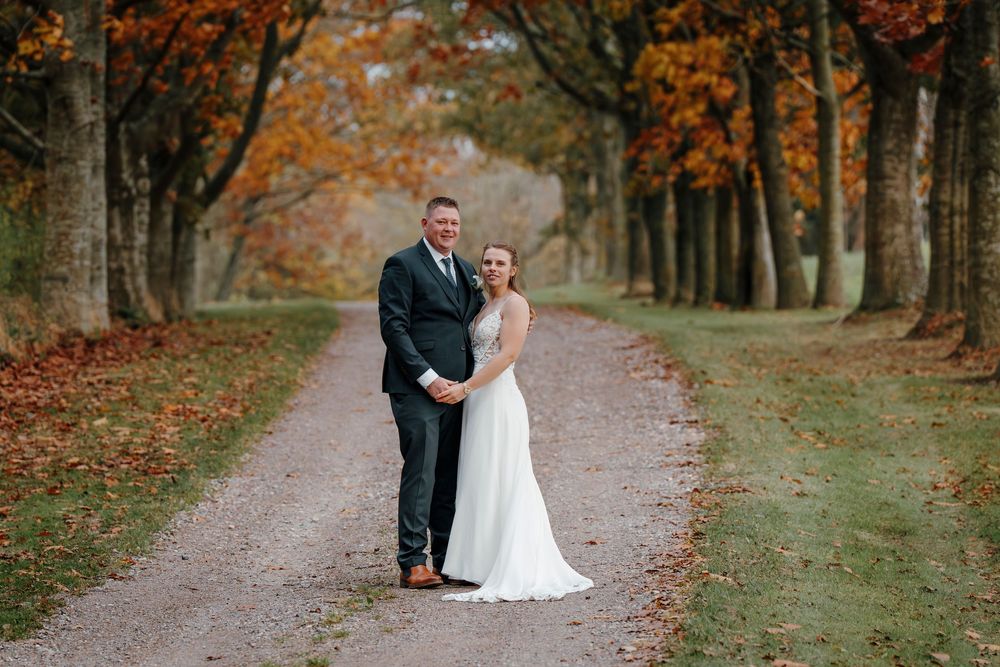 A couple poses together on a tree-lined autumn path with orange and red fall foliage creating a romantic backdrop.