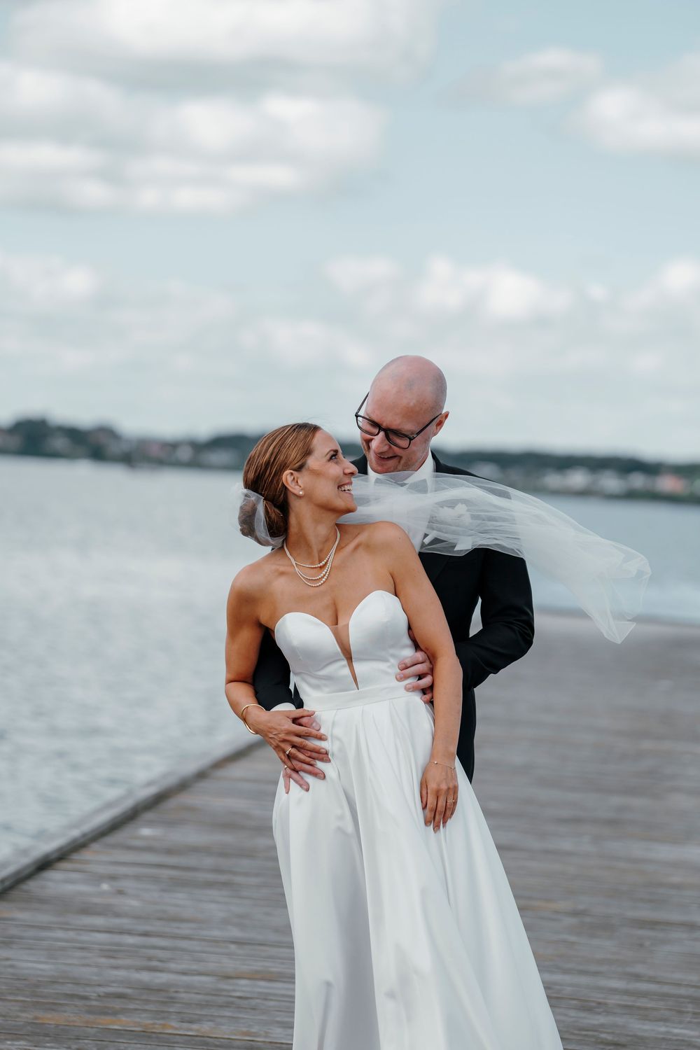 Bride and groom embrace on a waterfront dock, her veil blowing in the wind, wearing a white gown and black suit.