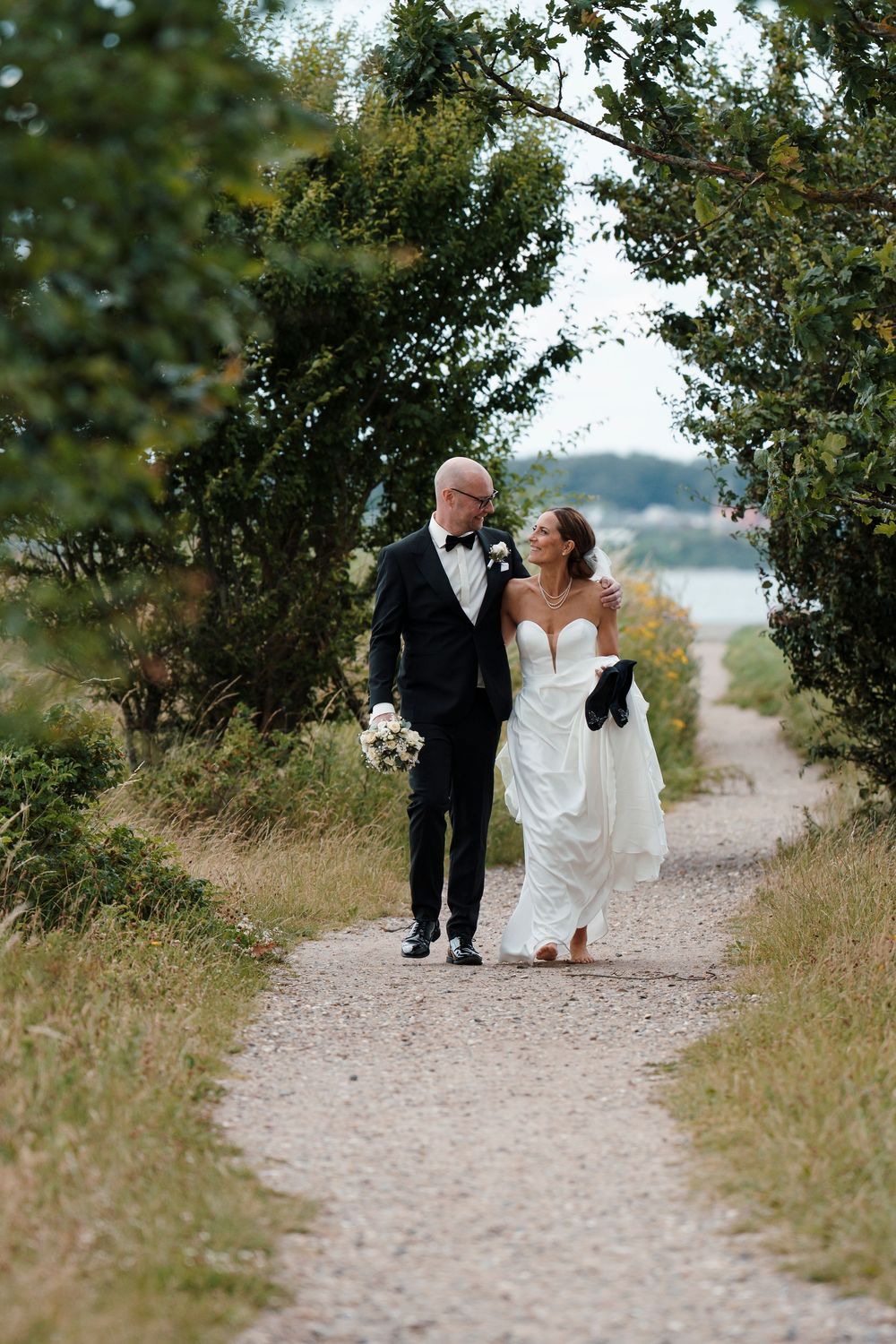 A bride and groom walk hand in hand along a scenic gravel path surrounded by lush greenery on their wedding day.
