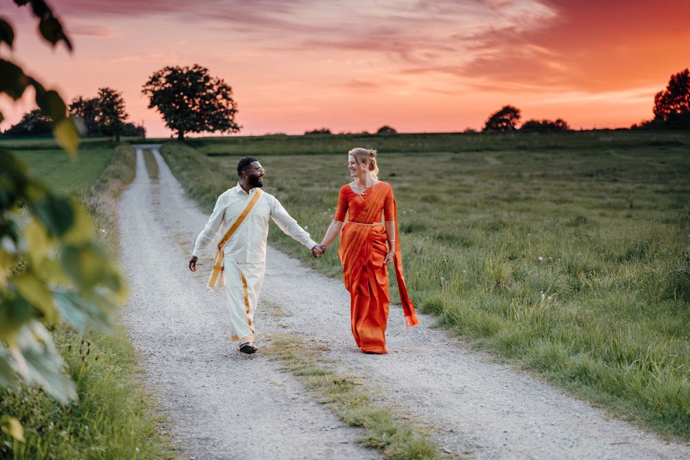 Couple in traditional Indian wedding attire walking hand-in-hand on a gravel path through green fields at sunset.