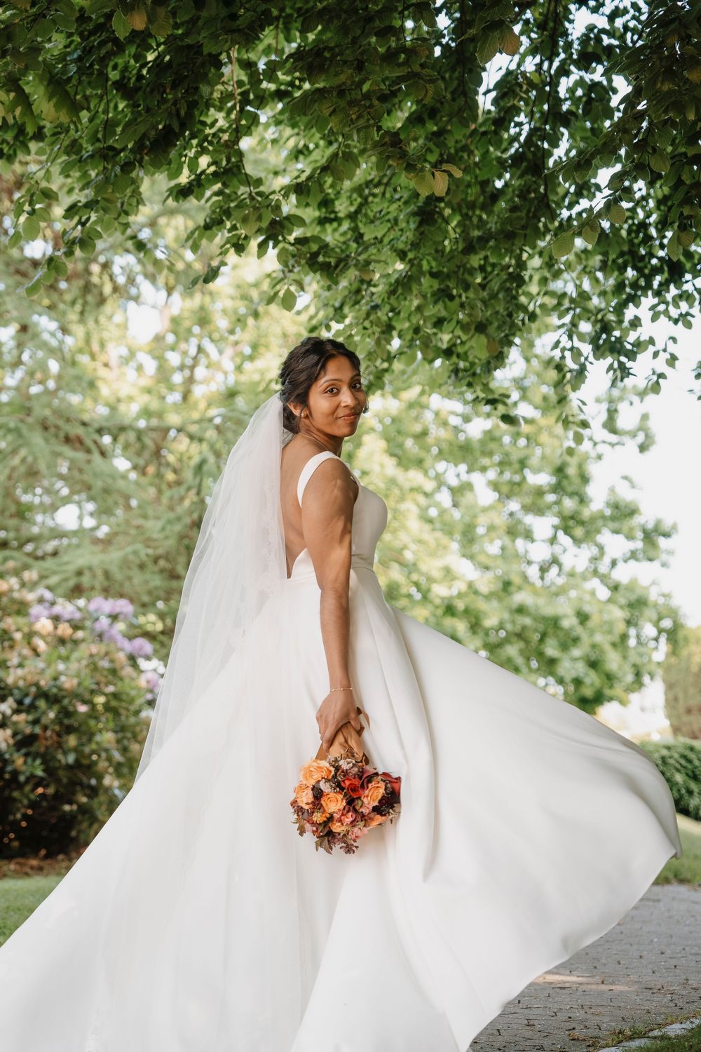 Bride in flowing white dress twirls outdoors holding orange bouquet under green trees.