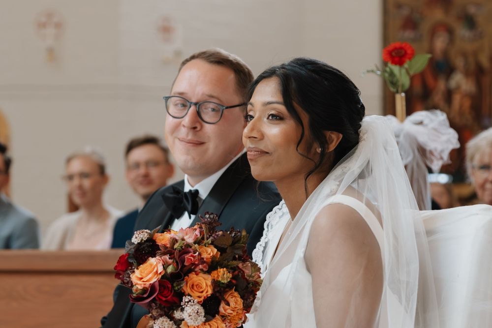 Couple in wedding attire sitting together during church ceremony with floral bouquet.