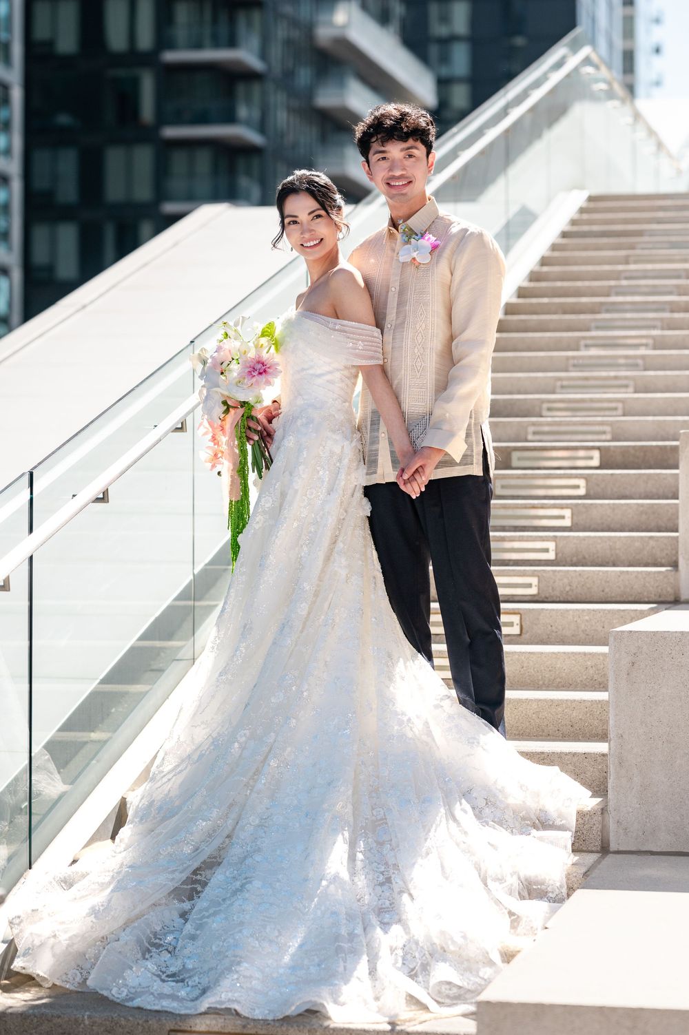 Bride in a lace gown holding a floral bouquet beside a groom in a barong tagalog on the outdoor stairs of a modern venue.