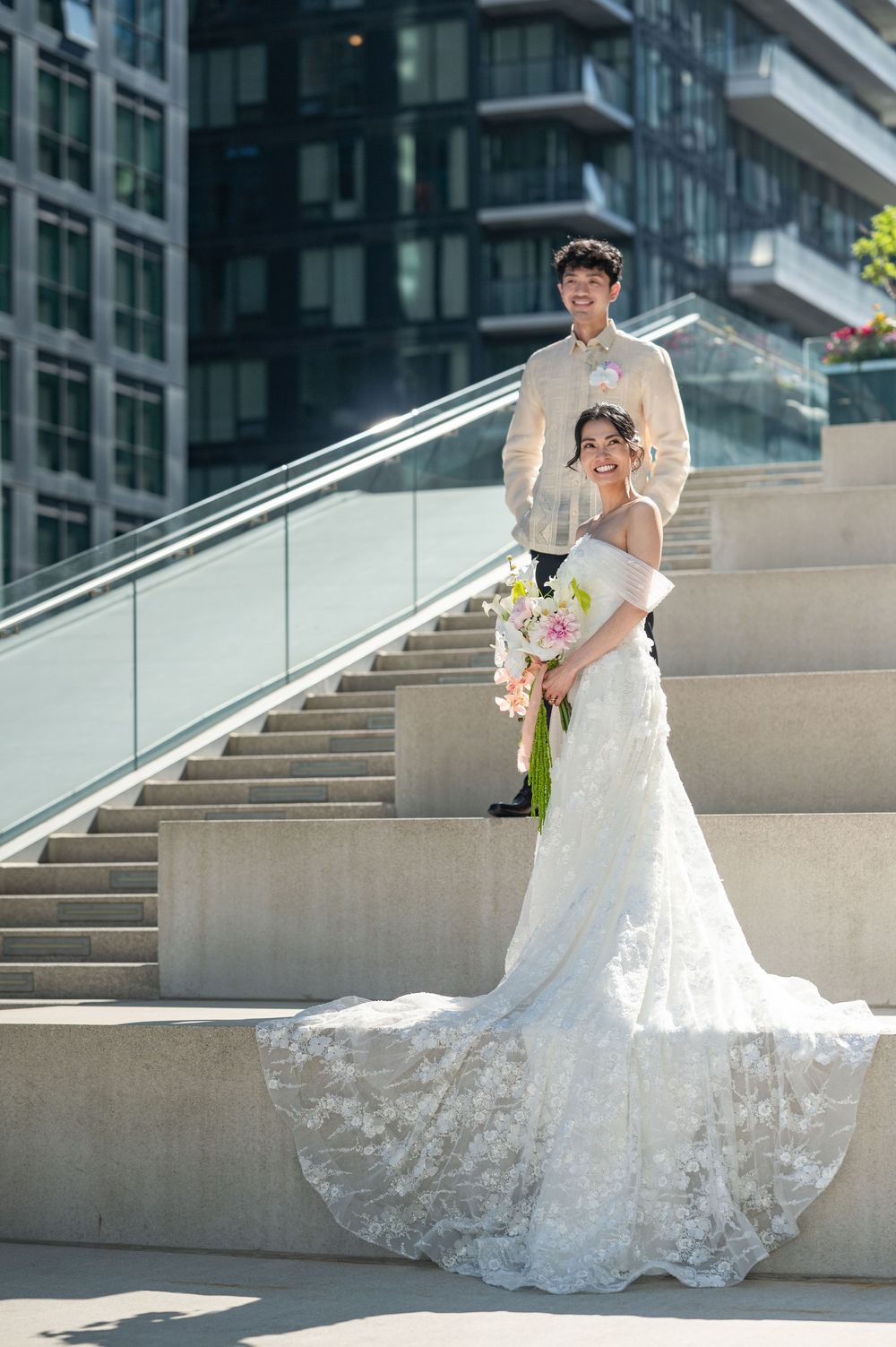 Bride in a white lace gown holding a bouquet on stone steps, groom in a beige suit standing behind her.