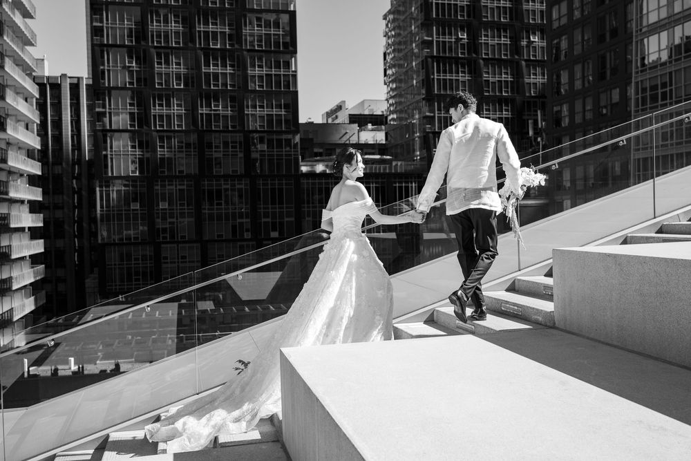 Black and white photo of a bride and groom holding hands walking up outdoor stairs with city buildings in the background.