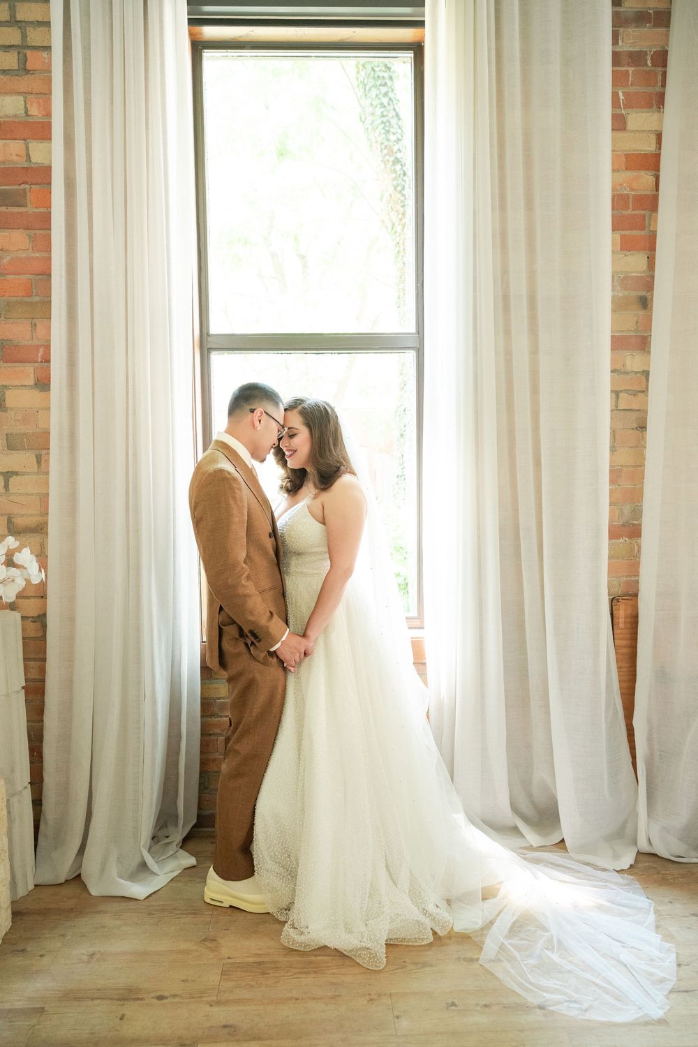 Bride and groom sharing a romantic moment by a sunlit window in a rustic brick venue on their wedding day.