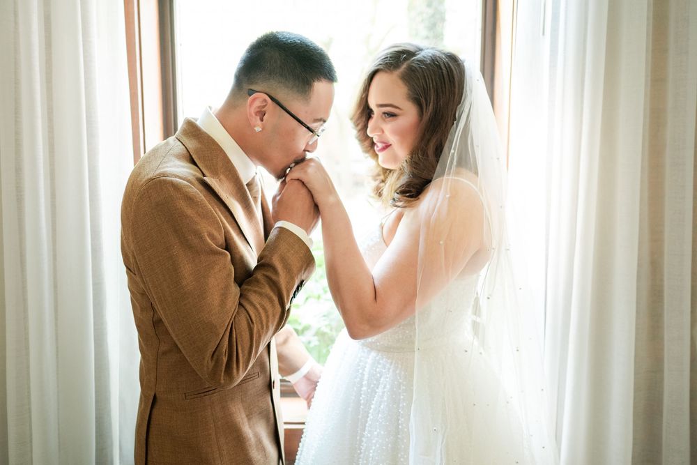 Groom in a tan suit kissing the bride's hand beside a sunlit window, her veil catching the light.
