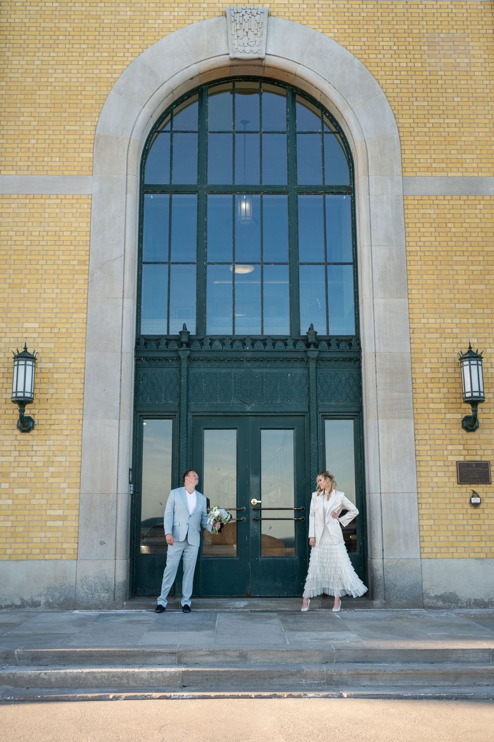 Bride and groom standing a few feet apart in front of a grand arched window in a yellow brick building.