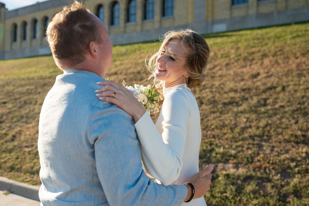 Bride and groom embracing outdoors on their wedding day, she holding white flowers, near a historic New York building.