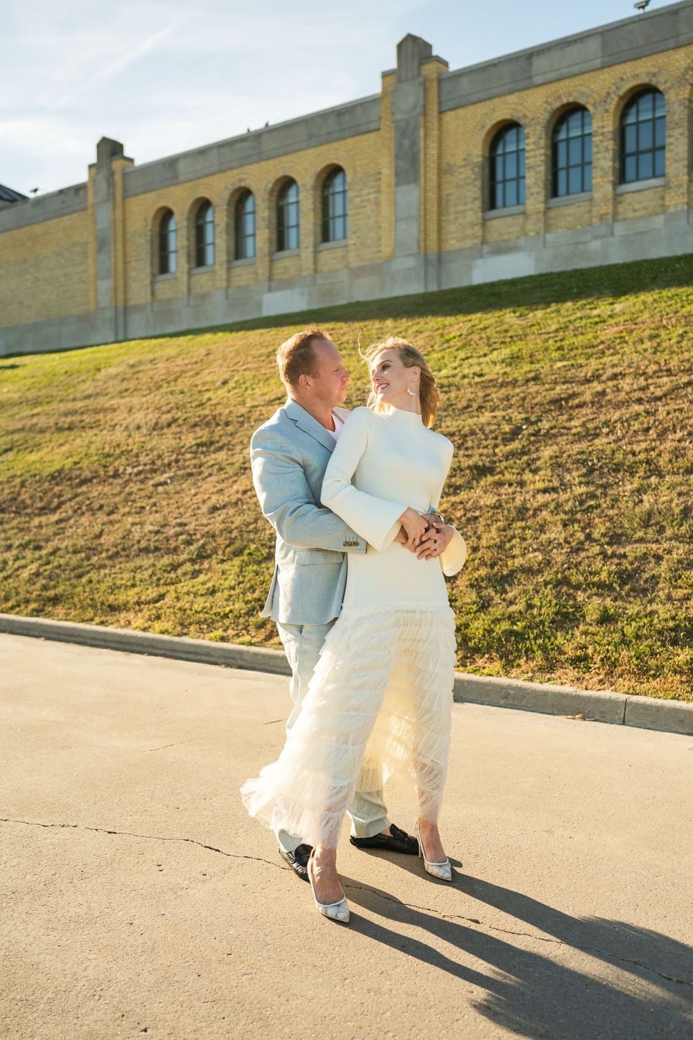 Bride and groom dancing outdoors beside a stone building in bright afternoon sun, she in white, he in a light blue suit.
