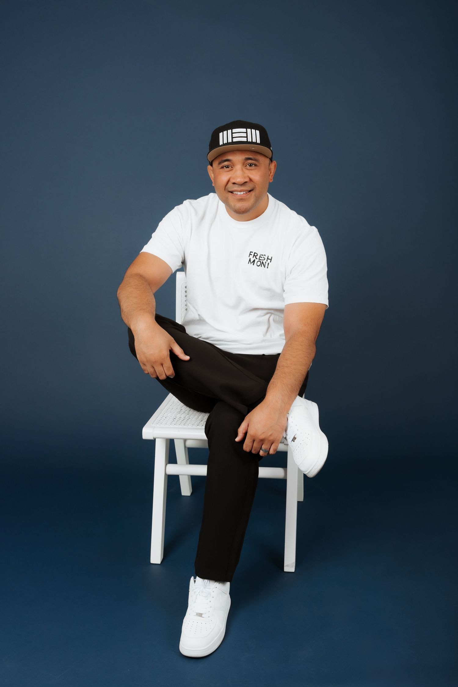Man in white Fresh Men t-shirt and black cap sits relaxed on white chair against navy blue background.