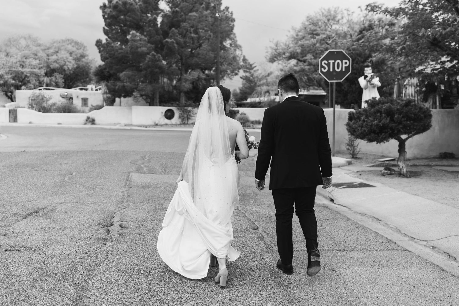 Black and white photo of a bride and groom walking hand in hand toward a stop sign on their wedding day.