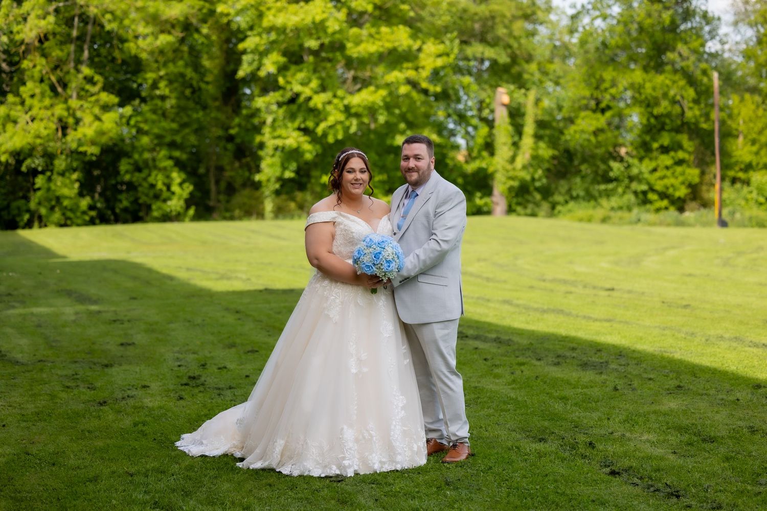 Bride in white gown holding blue bouquet stands with groom in gray suit on lush green lawn. Wedding at The Story, Durham. Casual Captures