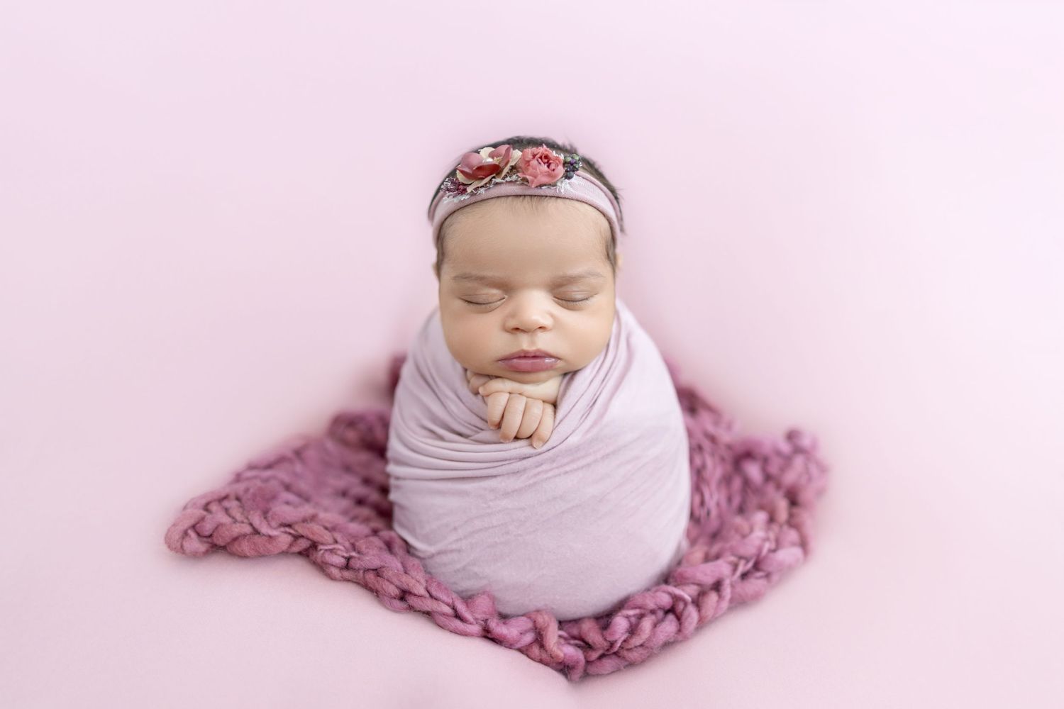 Sleeping newborn baby swaddled in lavender wrap with floral headband on pink background.