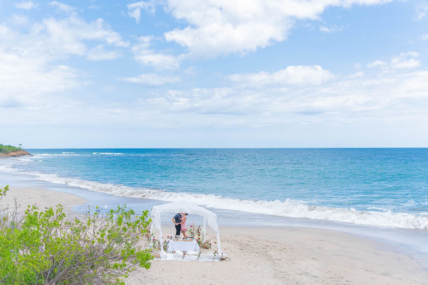 Couple stands under a white canopy on a serene tropical beach with turquoise ocean waves and lush greenery nearby.