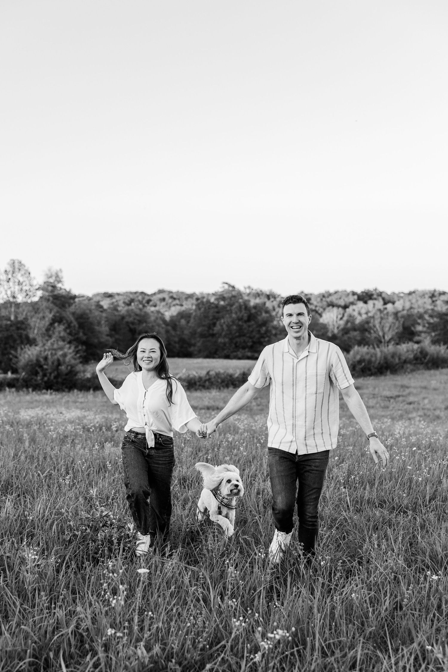 Black and white photo of a happy couple holding hands and walking with their dog through a scenic meadow.