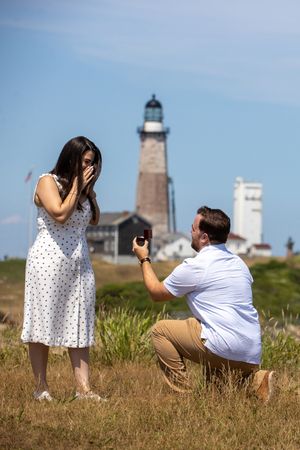 Man proposing on one knee to surprised woman in white polka dot dress near historic lighthouse.