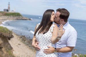 Couple sharing a kiss after proposal with lighthouse and ocean visible in the background.