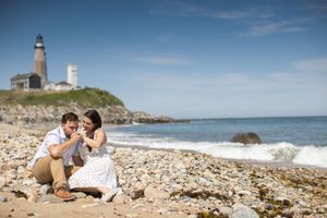 Newly engaged couple sitting on rocky beach admiring engagement ring with lighthouse backdrop.