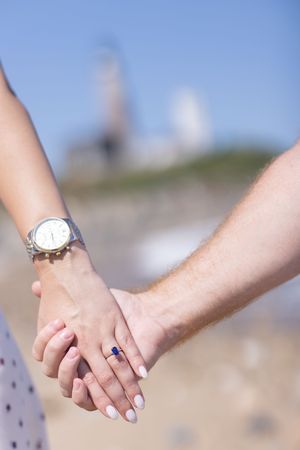 Close-up of couple holding hands showing sapphire engagement ring and silver watch near lighthouse.