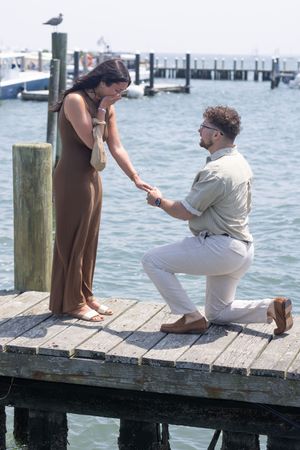 Man proposing on one knee on a wooden dock to woman in brown dress at a sunny marina.