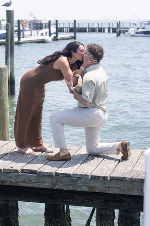 Couple sharing a kiss during a marriage proposal on a waterfront dock with marina in background.