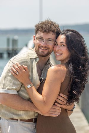 Happy newly engaged couple embracing and smiling together on a bright sunny waterfront pier.