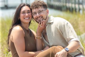 Smiling engaged couple sitting closely together outdoors showing off engagement ring in sunlight.
