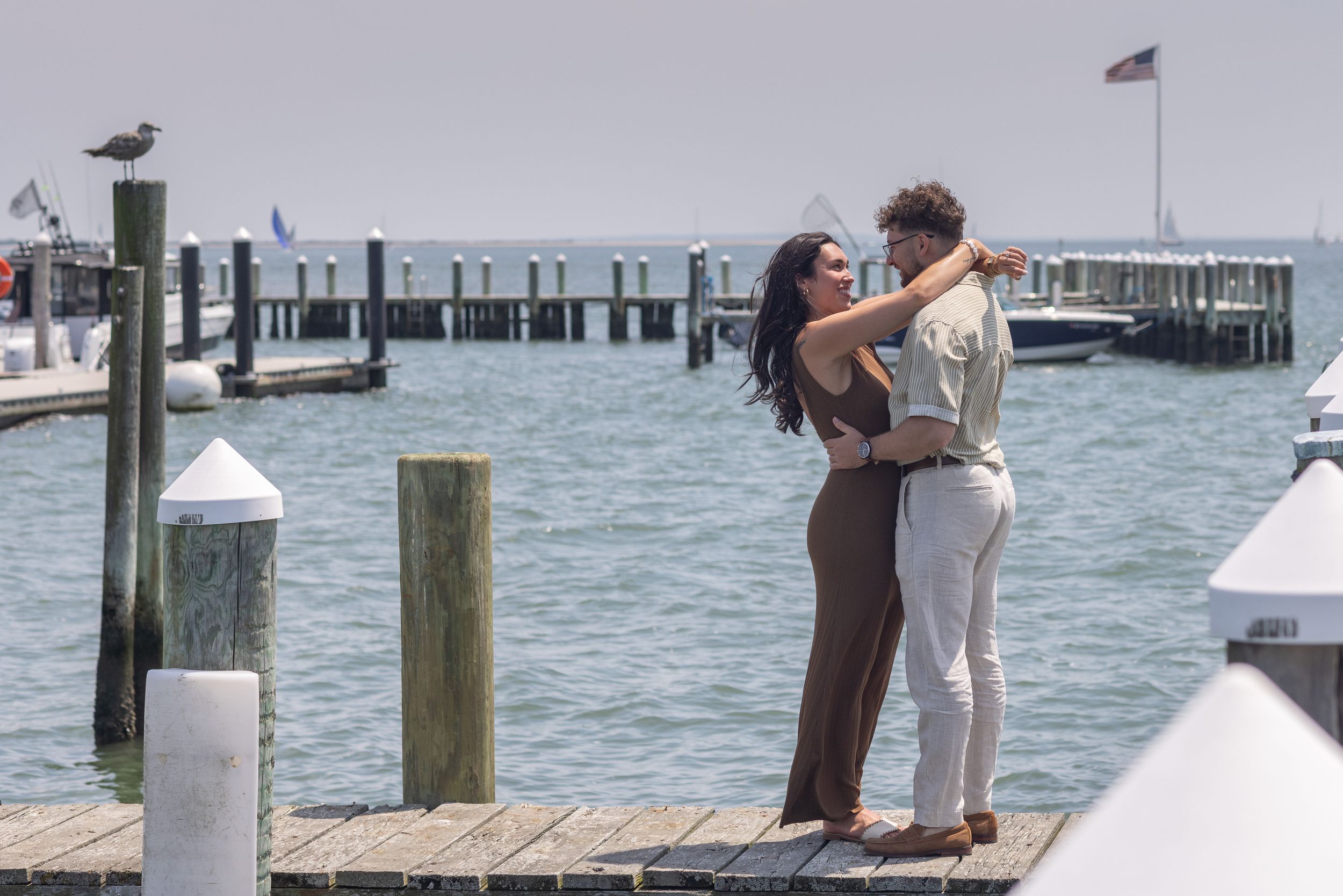 Newly engaged couple hugging romantically on a wooden dock overlooking a scenic marina waterway.