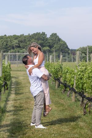 Man lifting woman joyfully in his arms between rows of grapevines at a sunny vineyard.