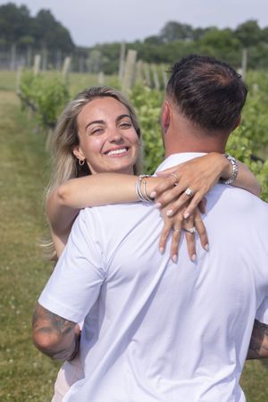 Smiling blonde woman embracing man from behind showing engagement ring in a green vineyard.
