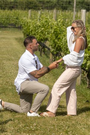 Man on one knee proposing to blonde woman with sunglasses in a sunny vineyard setting.