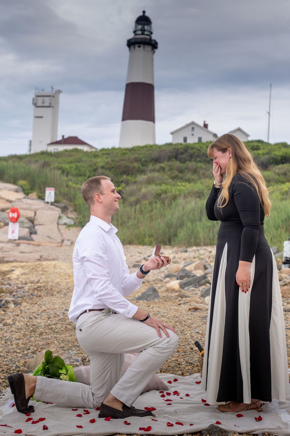Man proposing on one knee to his surprised girlfriend at a scenic lighthouse beach with rose petals.
