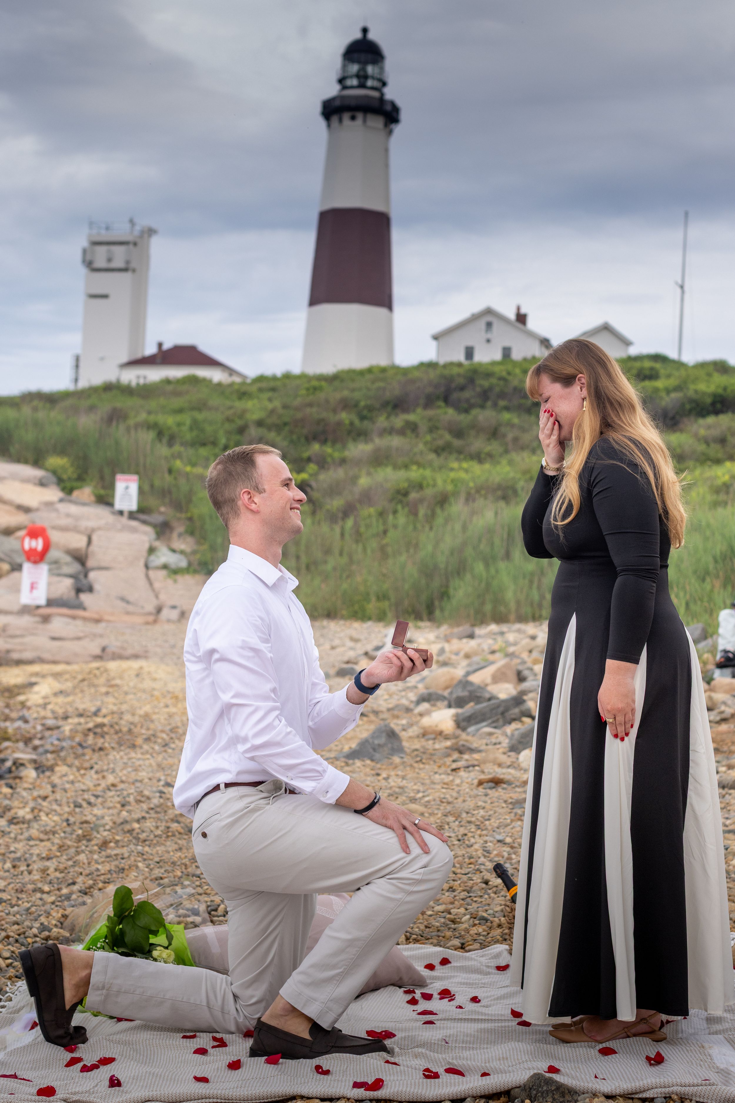 Man proposing on one knee to his surprised girlfriend at a scenic lighthouse beach with rose petals.