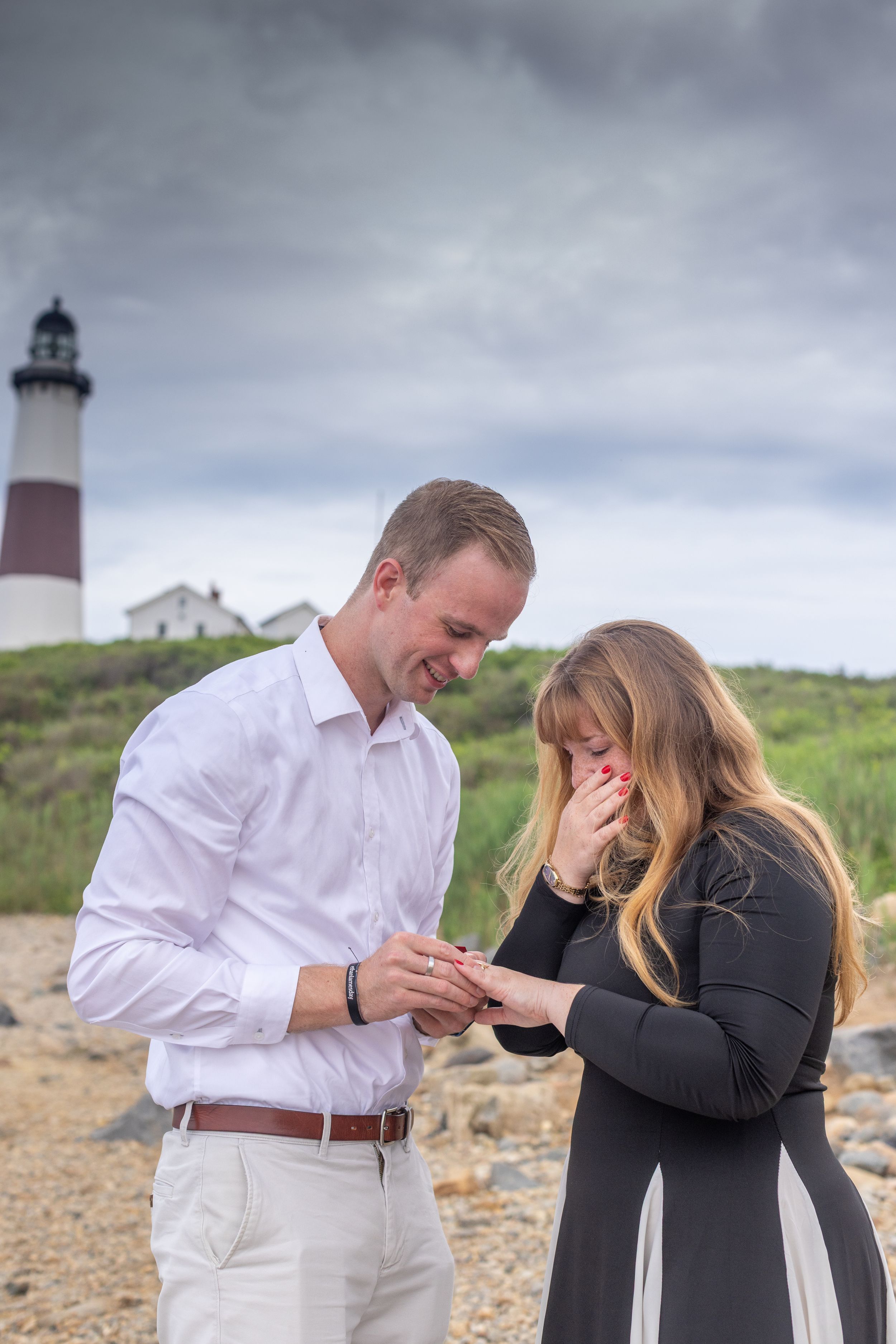 Emotional couple sharing a ring placement moment during a romantic lighthouse beach proposal.