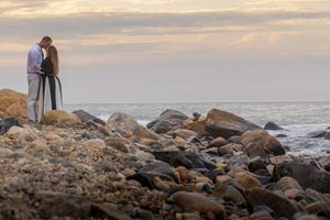 Romantic couple embracing on a rocky beach at sunset with ocean waves in the background.